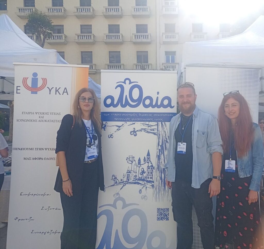 Ομαδική φωτογραφία μπροστά σε ενημερωτικά banner υπαίθριας δράσης ψυχικής υγείας.