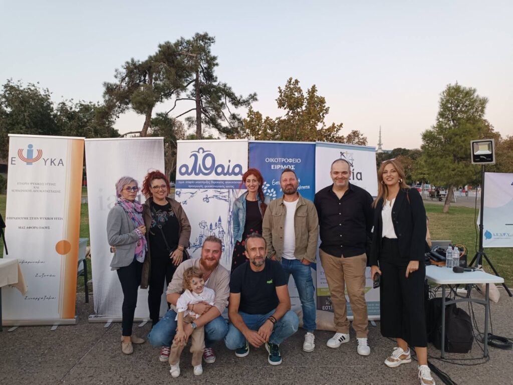 Ομαδική φωτογραφία συμμετεχόντων μπροστά σε banner της ΑΛΘΑΙΑ σε υπαίθριο φεστιβάλ.