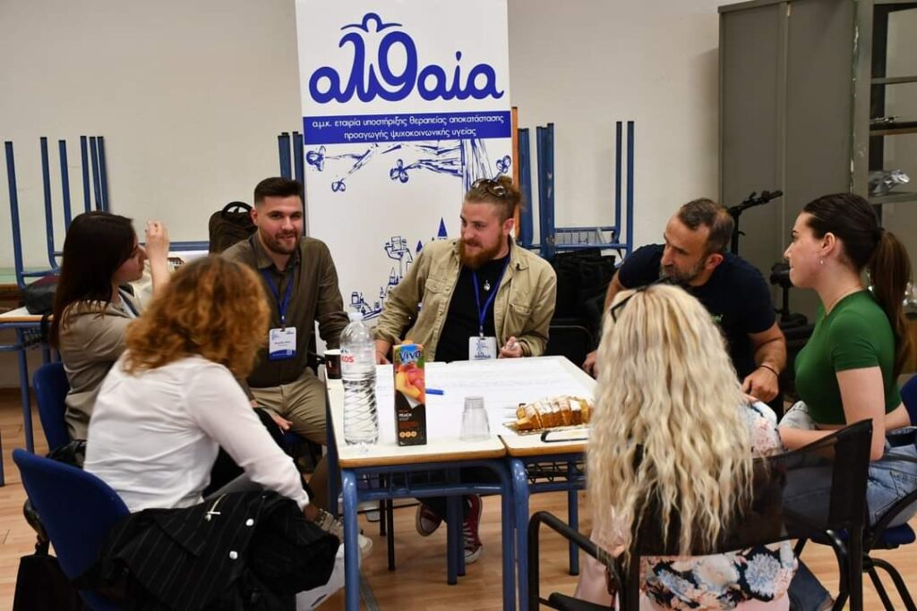 Συμμετέχοντες συζητούν γύρω από τραπέζι σε εργαστήριο, μπροστά σε banner της ΑΛΘΑΙΑ.